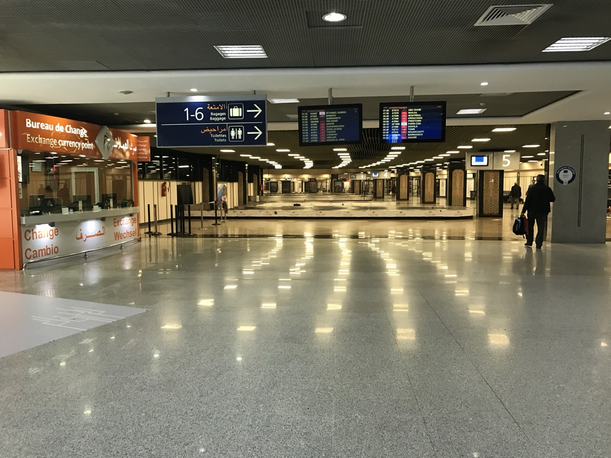 An empty baggage carousel in an airport arrivals hall with a few travelers waiting nearby.