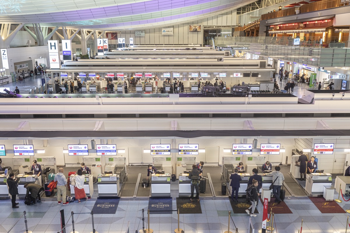 Travelers with suitcases queuing at airline check-in counters in a bright Japanese airport terminal.
