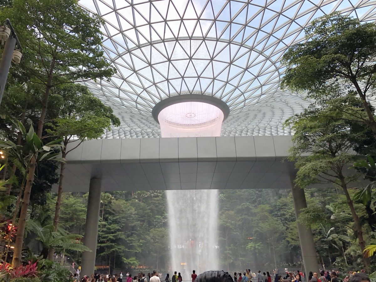 The Rain Vortex indoor waterfall pouring through the glass dome of Jewel Changi Airport, surrounded by terraced indoor forest.
